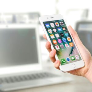 Close-up view of a smartphone displaying apps, held by a hand, with a blurred laptop in the background.